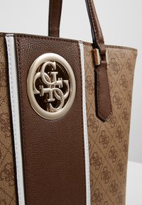 Brązowa torba tote z teksturowanego materiału, z białymi i złotymi akcentami, dużym metalowym logo oraz usztywnionym kształtem z podwójnymi uchwytami.