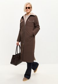 Brun lang frakke med lynlås foran, pelskrave, to brystlommer og sidelommer. Kombineret med mørke jeans og taupe sko.