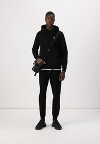Svart hettegenser med snøring, svarte bukser og svarte joggesko. Modellen holder en liten svart veske over kroppen. Enkelt design og ensfargede farger.