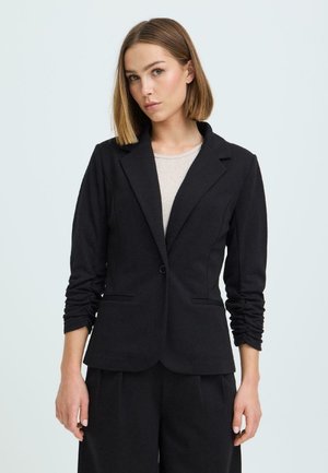 Kobieta o prostych włosach do ramion, ubrana w czarną marynarkę zapinaną na guziki i czarne spodnie, na białym tle.