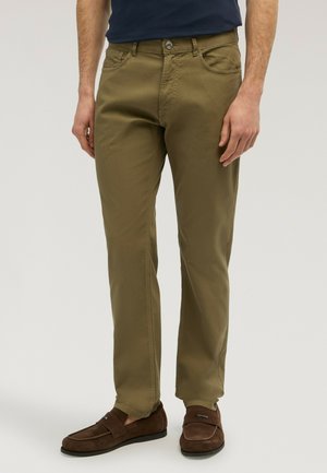 Homme portant un pantalon droit olive et des mocassins en daim marron, debout devant un fond clair uni.