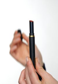 Svart leppestift-tube med gullakenter, lett avsmalnende, holdt av hender. Kappen fjernet, og avslører en rødlig leppestiftspiss. Glatt overflate.