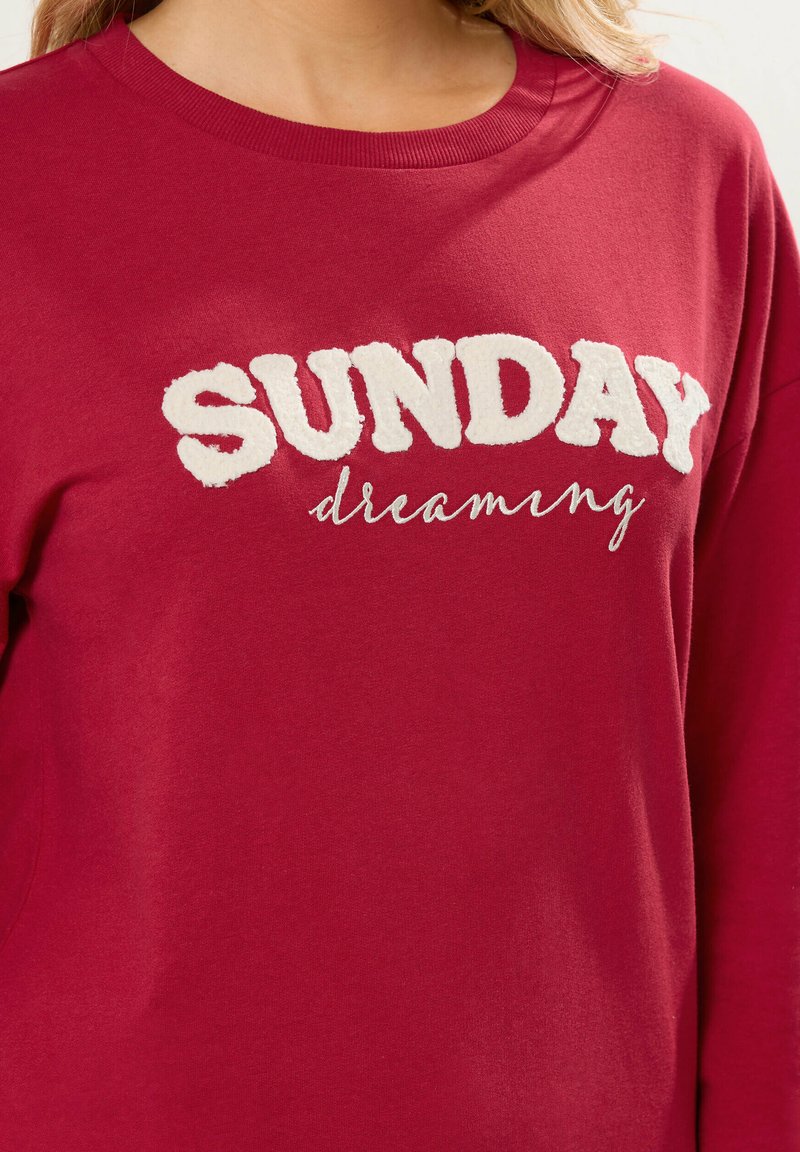 Sudadera burdeos con un texto bordado en blanco elevado que dice "SUNDAY" y "dreaming" en un estilo cursivo abajo. Textura suave.