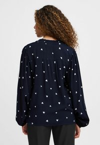 Blouse marine avec imprimé cœurs blancs, manches longues, épaules froncées et coupe décontractée. Tissu doux avec une finition texturée.