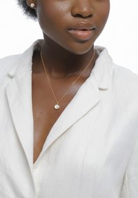 Guldhalskæde med en enkelt rund perle som vedhæng, båret sammen med en hvid linnedskjorte med krave, der fremhæver glatte teksturer og neutrale farver.