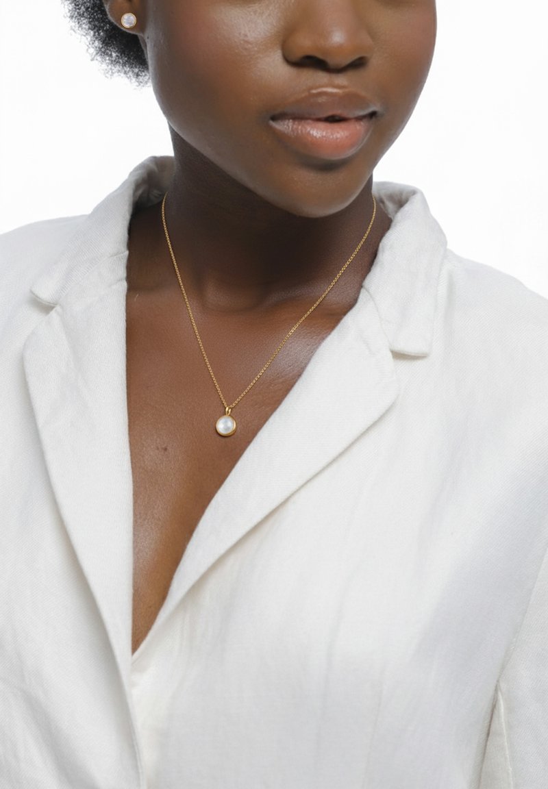 Guldhalskæde med en enkelt rund perle som vedhæng, båret sammen med en hvid linnedskjorte med krave, der fremhæver glatte teksturer og neutrale farver.