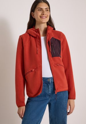 Kvinde iført rød fleecejakke med lynlås, hætte og brystlomme, blå jeans og en hvid t-shirt, smilende og med blikket let mod højre.