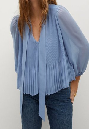 Mujer vistiendo una blusa plisada azul claro con mangas largas transparentes y una corbata, combinada con vaqueros azul oscuro.