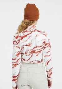 Langärmliges Oberteil mit weißem Grund und rotem Marmor-Muster, kombiniert mit hellbeigen Hosen. Das Modell trägt eine braune Strickmütze.