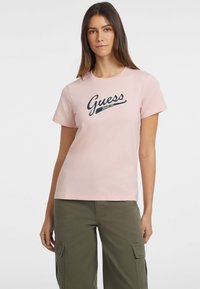 Jasnoróżowa koszulka z krótkim rękawem z granatowym logo "Guess Jeans 1981". Wykonana z bawełny, charakteryzuje się okrągłym dekoltem i luźnym krojem.