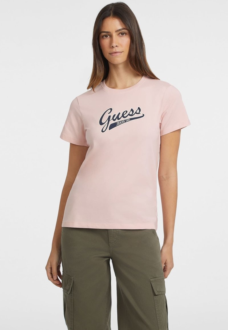 Jasnoróżowa koszulka z krótkim rękawem z granatowym logo "Guess Jeans 1981". Wykonana z bawełny, charakteryzuje się okrągłym dekoltem i luźnym krojem.