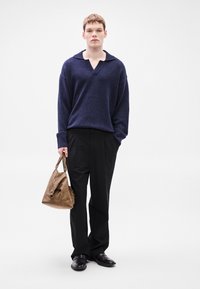 Μπλε σκούρο πλεκτό πουλόβερ με φαρδιά γιακά, σε συνδυασμό με μαύρο παντελόνι και γυαλιστερά μαύρα παπούτσια. Κρατάει καφέ δερμάτινη τσάντα χειρός με καμπύλο σχήμα.