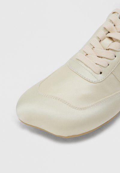 Αθλητικό παπούτσι σε κρεμ χρώμα με υφασμάτινα κορδόνια και ραμμένες λεπτομέρειες, εμφανιζόμενο από γωνία μπροστά-αριστερά σε λευκό φόντο.