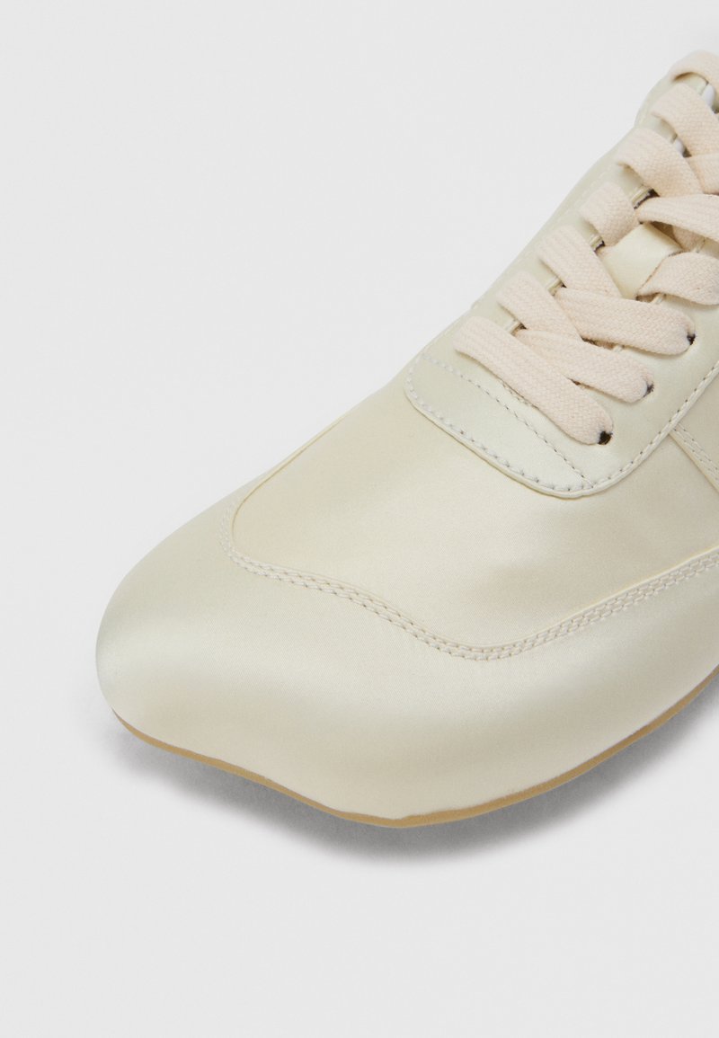 Sneaker i cremefarve med snørebånd af stof og syningdetaljer, vist fra front-venstre vinkel på en hvid baggrund.