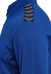 T-shirt de sport bleu à manches longues avec un tissu texturé, présentant un accent noir détaillé à l'épaule et un col haut.