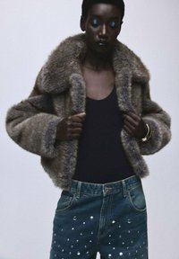 Chaqueta de piel sintética en gris-marrón con un amplio cuello, combinada con unos vaqueros azul oscuro que tienen tachuelas metálicas en la parte frontal. Top negro debajo.
