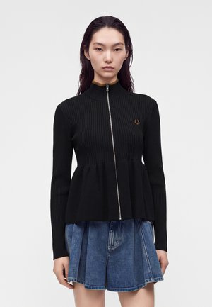 Woman with long dark hair wears a black zip-up ribbed sweater with a peplum hem and blue pleated denim shorts against a plain background.