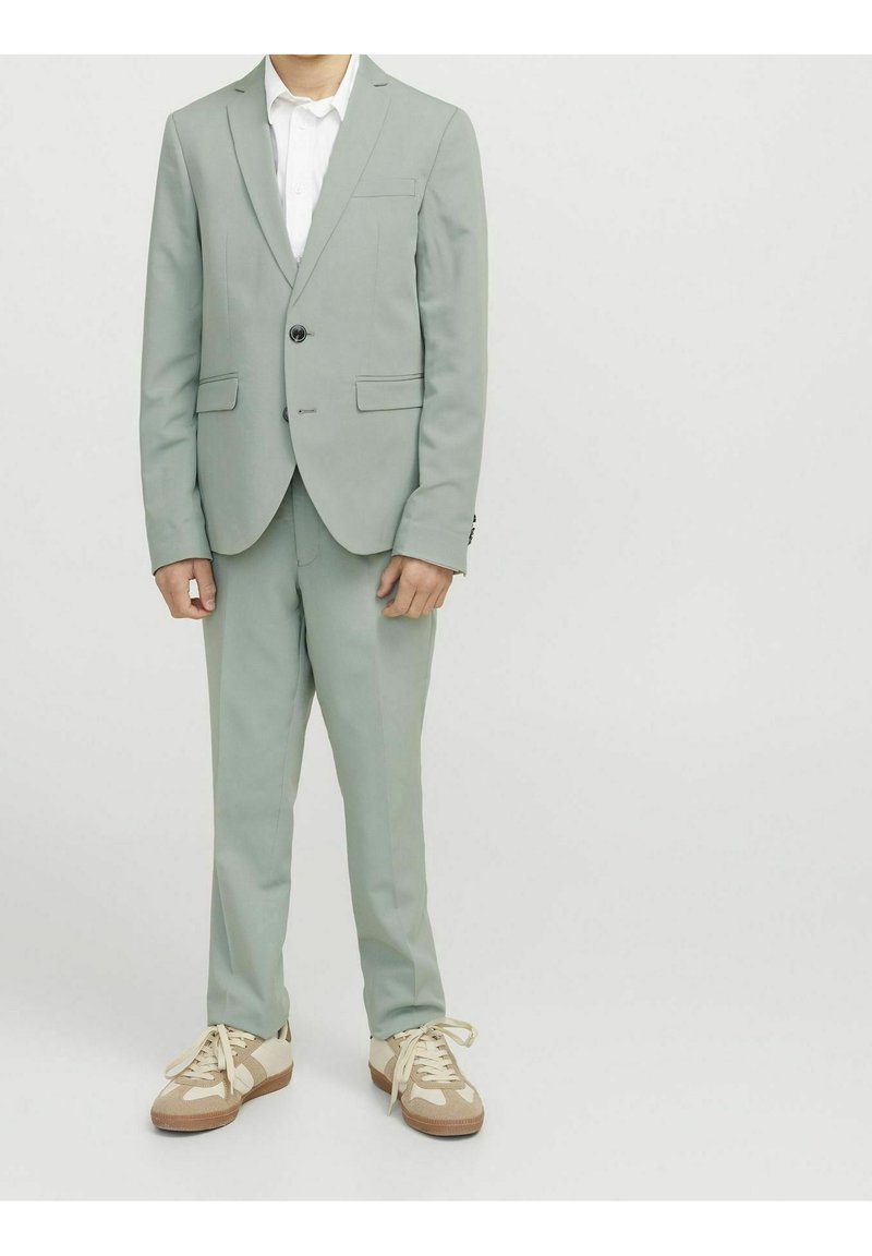 Jasnozielony garnitur z jedno guzikową marynarką, dopasowanymi spodniami i białą koszulą. Uzupełniony brązowymi i kremowymi sneakerami.