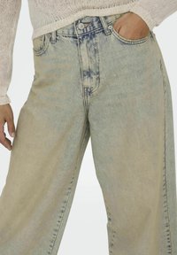 Jasnoniebieskie dżinsy w szerokim kroju wykonane z wyblakłego denimu, z postrzępionymi krawędziami, wysokim stanem i pięcioma kieszeniami. Model częściowo widoczny.