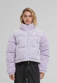 Lilla puffjakke med høy krage, vertikal ribbet tekstur og glidelås. Har et brodert logo på brystet. Ledig passform.