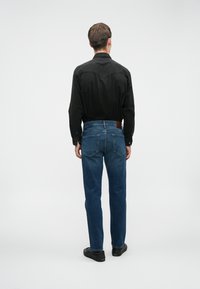Svart skjorte med western-stil yoke, sammen med blå denimjeans. Mørke sko fullfører antrekket. Ensfarget bakgrunn, full baksidevisning.