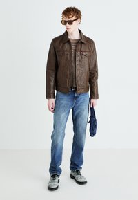 Braune Lederjacke mit knöpfbarem Vorderteil und zwei Brusttaschen, getragen über einem Strickpullover; kombiniert mit hellblauen Jeans und grauen Schuhen.