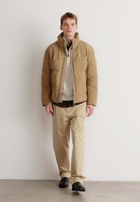Tan quilted pufferjakke med høj krave, båret over en cremefarvet sweater og en mørk skjorte, parret med beige bukser og sorte sko.