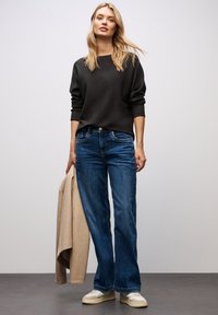 Maglione nero a maniche lunghe abbinato a jeans svasati blu. Giacca beige tenuta in una mano, scarpe da ginnastica ai piedi. Design semplice e minimale.