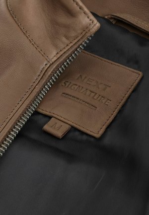 Giubbotto di pelle marrone con zip argento. L'etichetta riporta "NEXT SIGNATURE," indicando la taglia M. Fodera in tessuto nero e dettagli testurizzati.