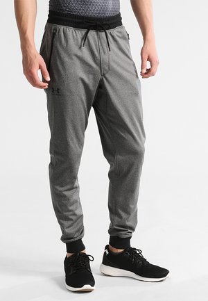 Homme portant un pantalon de jogging gris Under Armour avec taille ajustable par cordon et des chaussures de sport noires sur fond blanc.