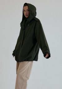 Przesadzony oliwkowy zielony płaszcz z kapturem, zapinany na guziki, z regulowanym ściągaczem na dole oraz kieszeniami po bokach. Gładka tekstura materiału.