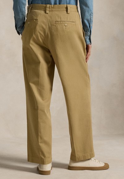 Vue de dos d'une personne portant un pantalon beige droit, des baskets blanches et une chemise bleue à manches longues, debout devant un fond neutre.