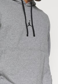 Hombre que lleva una sudadera gris con cordones negros y un logo de baloncesto en silueta negra en el pecho.