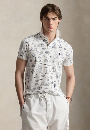 Ung mand iført hvid poloshirt med nautisk-tema skitser og hvide bukser, stående mod en ensfarvet beige baggrund.