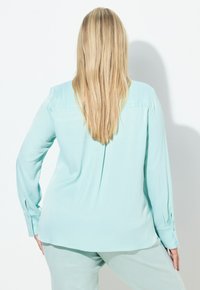 Lyse turkis bluse med lange ærmer, knapmanchetter, en læg bagpå og glat tekstur. Kombineret med matchende lyse bukser.