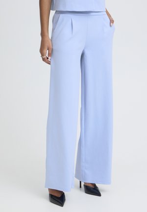 Femeie purtând pantaloni largi albastru deschis și tocuri negre ascuțite, cu o mână în buzunar și ojă albă vizibilă.