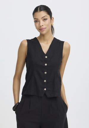 Sötét hajú nő, akinek hátra kötött haját fekete ujjatlan, gombos felső és fekete nadrág egészíti ki, kezét zsebébe dugva, arany kerek fülbevalókkal.