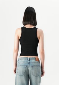Frau mit glattem schwarzen Haar, die ein ärmelloses schwarzes Oberteil und hellblaue Jeans trägt, steht vor einem weißen Hintergrund.