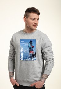 Uomo che indossa una felpa grigia con grafica da alpinista, bussola e testo "Since 1981" e "Durham Mountaineering", con le mani nelle tasche.