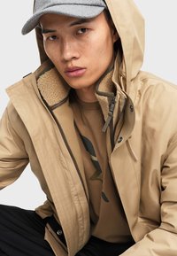 Beige Jacke mit strukturiertem Innenfutter, Reißverschluss und Druckknopfverschlüssen sowie einer abnehmbaren Kapuze. Getragen über einem sandfarbenen Shirt mit einem grünen Grafikdesign.