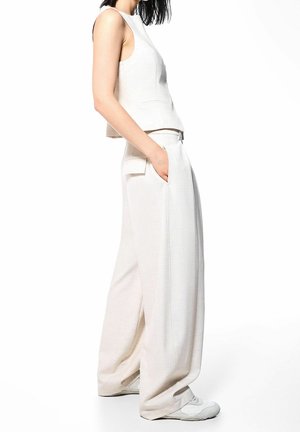 Beige cropped sleeveless top with a high neckline, paired with loose beige trousers featuring side pockets and a textured fabric.