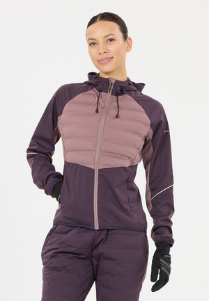 Frau trägt eine lilafarbene, wattierte Jacke mit Kapuze und passende Hose, posiert mit einer Hand in der Tasche und trägt schwarze Handschuhe.