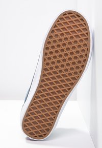 Semelle d’une basket décontractée avec une semelle extérieure en caoutchouc marron à motif gaufré, une semelle intermédiaire blanche et une tige bleue, positionnée verticalement sur fond blanc.