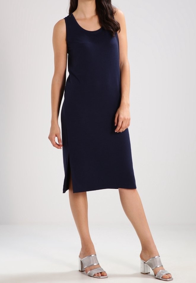 Femme portant une robe midi bleu marine sans manches avec une fente latérale, associée à des sandales argentées à talon bloc et bout ouvert, debout sur un sol blanc.