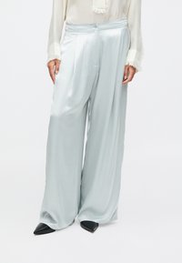 Světle modré saténové kalhoty širokého střihu s hladkou texturou, s puky na přední straně a knoflíkovým zapínáním, spárované s černými špičatými podpatky.