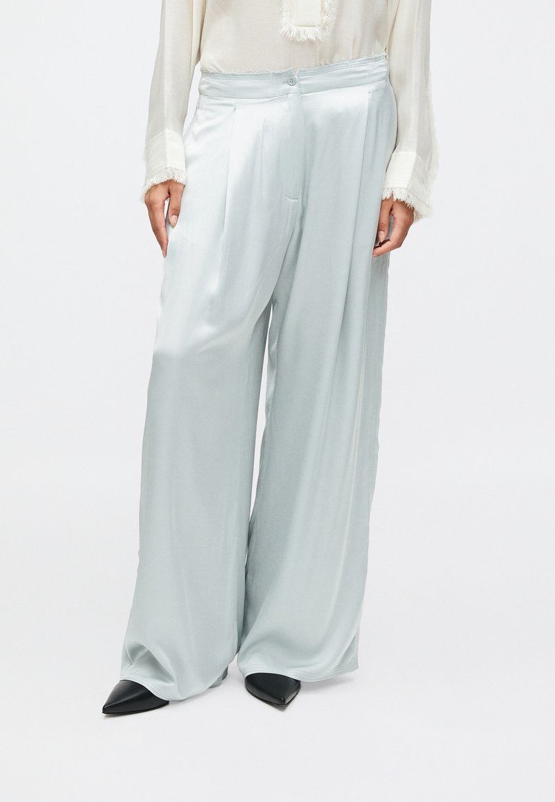 Světle modré saténové kalhoty širokého střihu s hladkou texturou, s puky na přední straně a knoflíkovým zapínáním, spárované s černými špičatými podpatky.