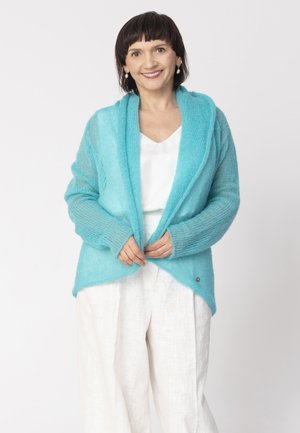 Kobieta z krótkimi ciemnymi włosami uśmiechająca się, ubrana w turkusowy otwarty kardigan na białej bluzce i luźnych białych spodniach, stojąca na jednolitym tle.