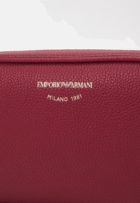 Ciemnoczerwona, teksturowana skóra z złotym logo Emporio Armani oraz wygrawerowanym napisem "Milano 1981" na powierzchni.