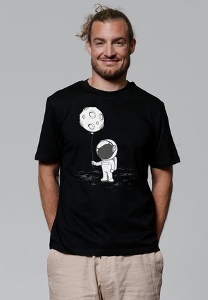 Zwart katoen t-shirt met een witte astronautafbeelding die een met patronen versierde maanballon vasthoudt, tegen een eenvoudige illustratie van een maanoppervlak.