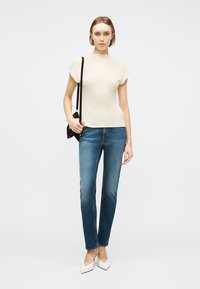 Kvinne iført kremfarget kortermet topp, blå jeans med rette ben, hvite spisse sko, og bærende på en liten svart skulderveske.